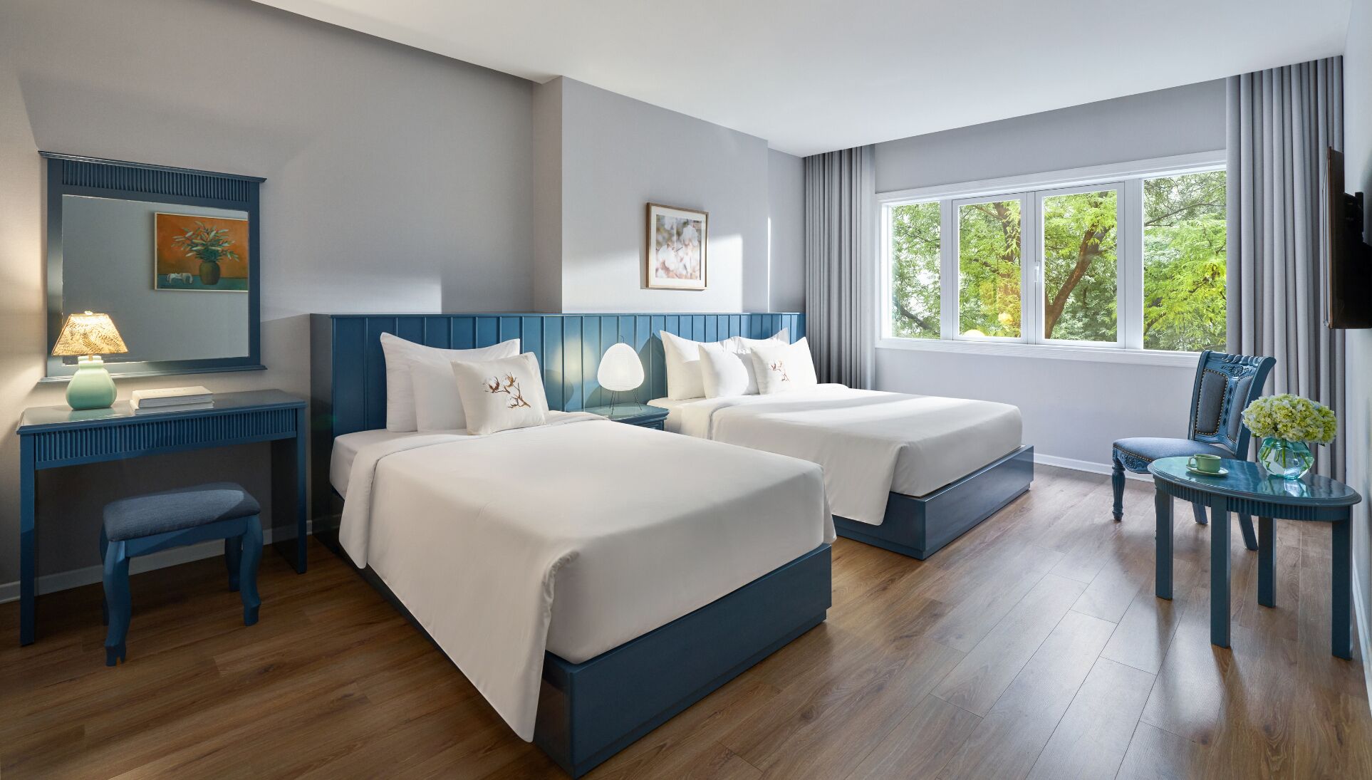 A hotel room with two beds, a desk, a chair, a TV, and a window with a view of trees.