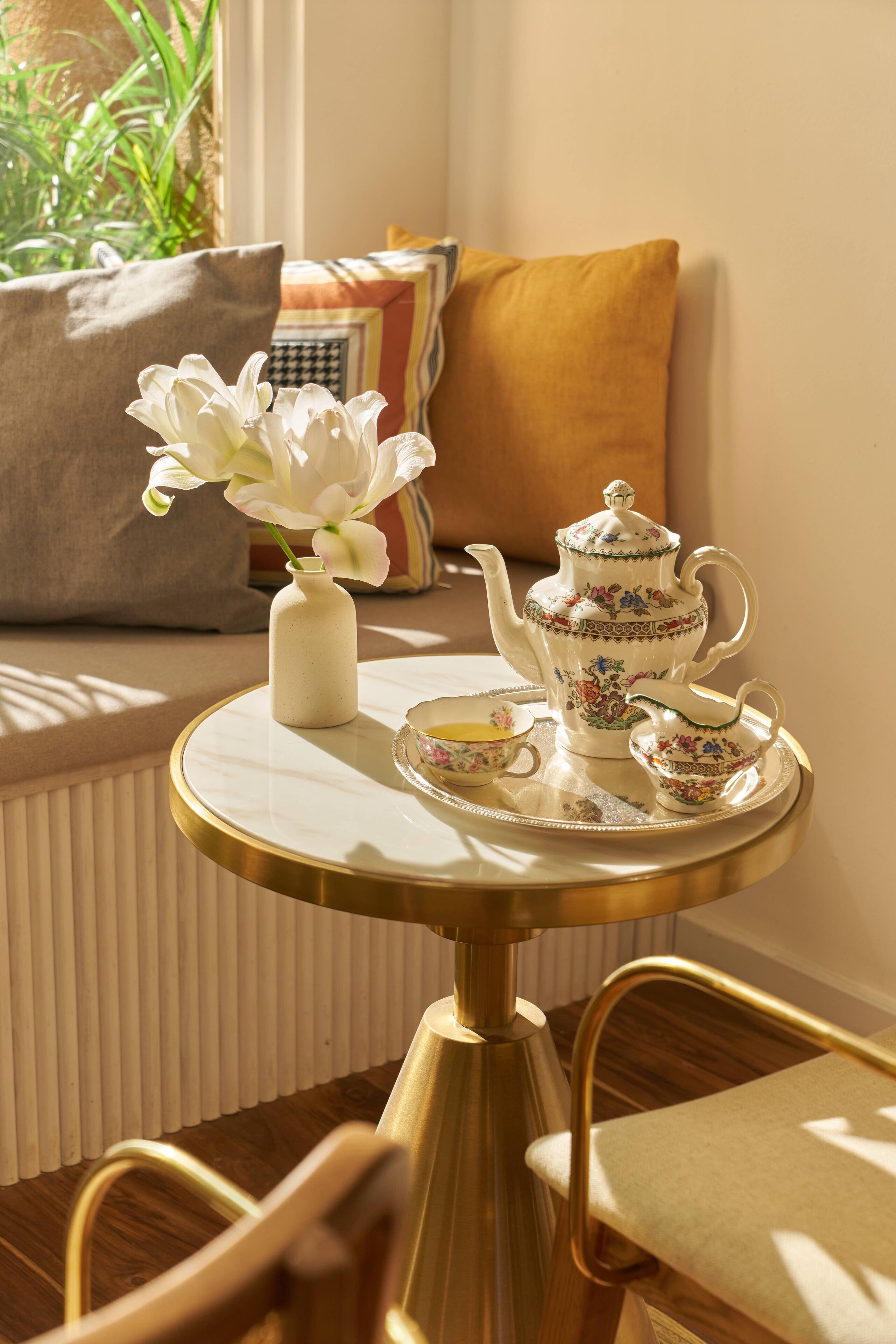 A table with a tea set, a vase with flowers, and a window with a view of greenery.