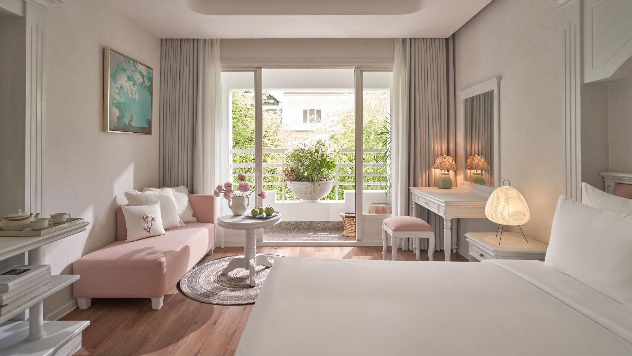 A hotel room with a bed, a sofa, a desk, a chair, a table with flowers, and a view of greenery outside.
