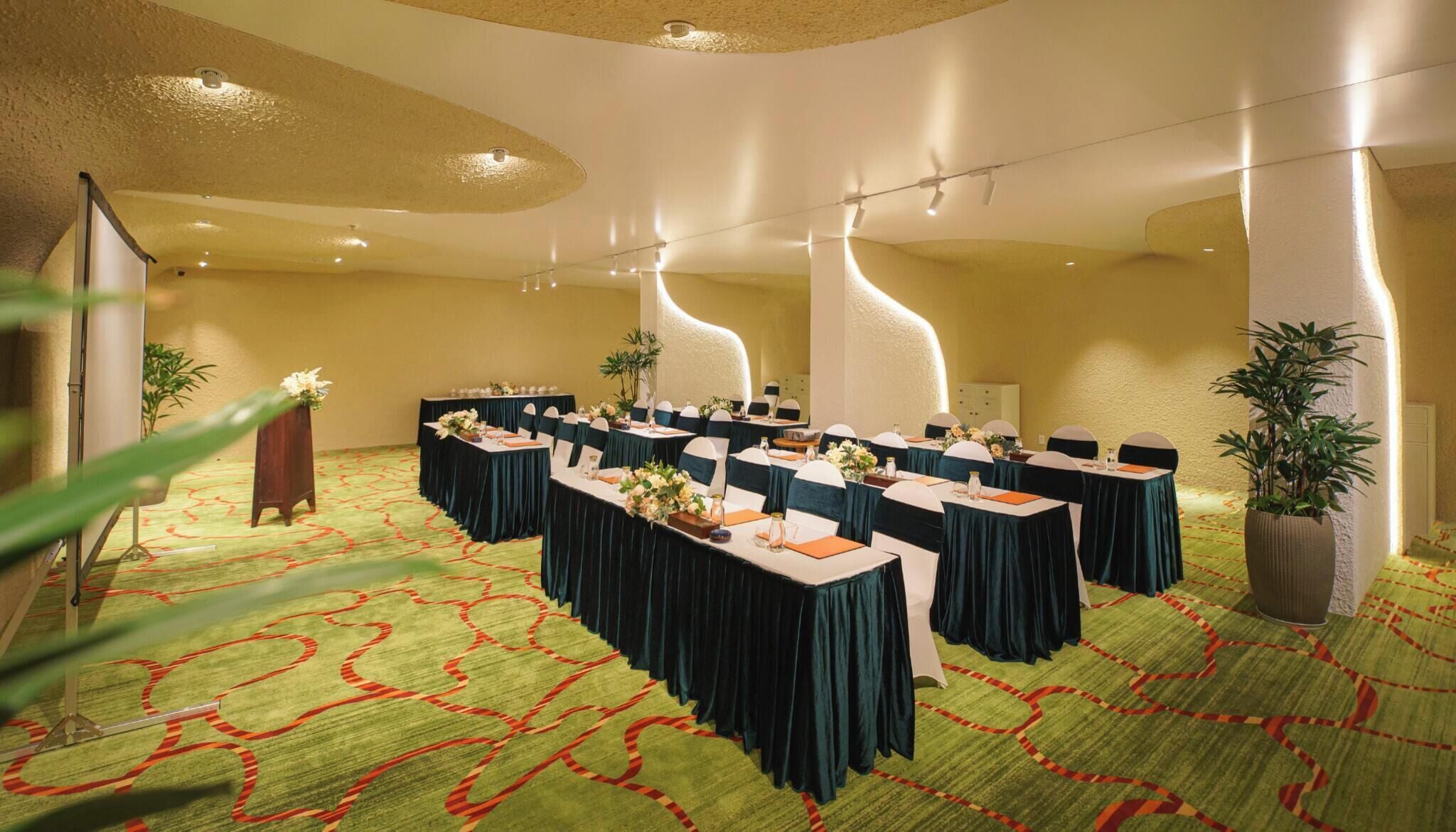 A conference room with tables set for a meeting, a large potted plant, and a carpeted floor.