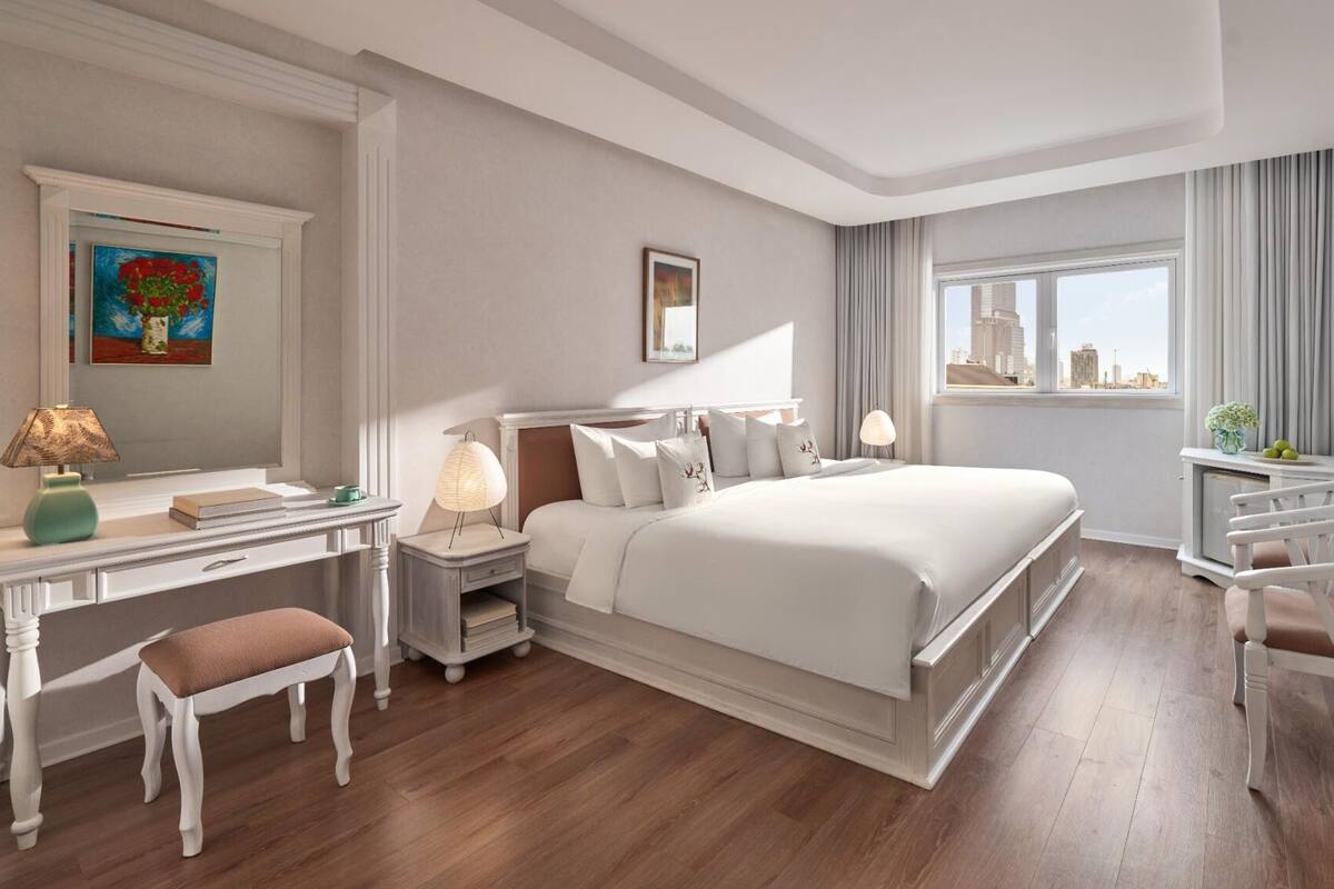Quarto de hotel com uma cama grande, uma escrivaninha com cadeira, mesa de cabeceira com abajures, TV fixada na parede e janela com vista para a cidade.