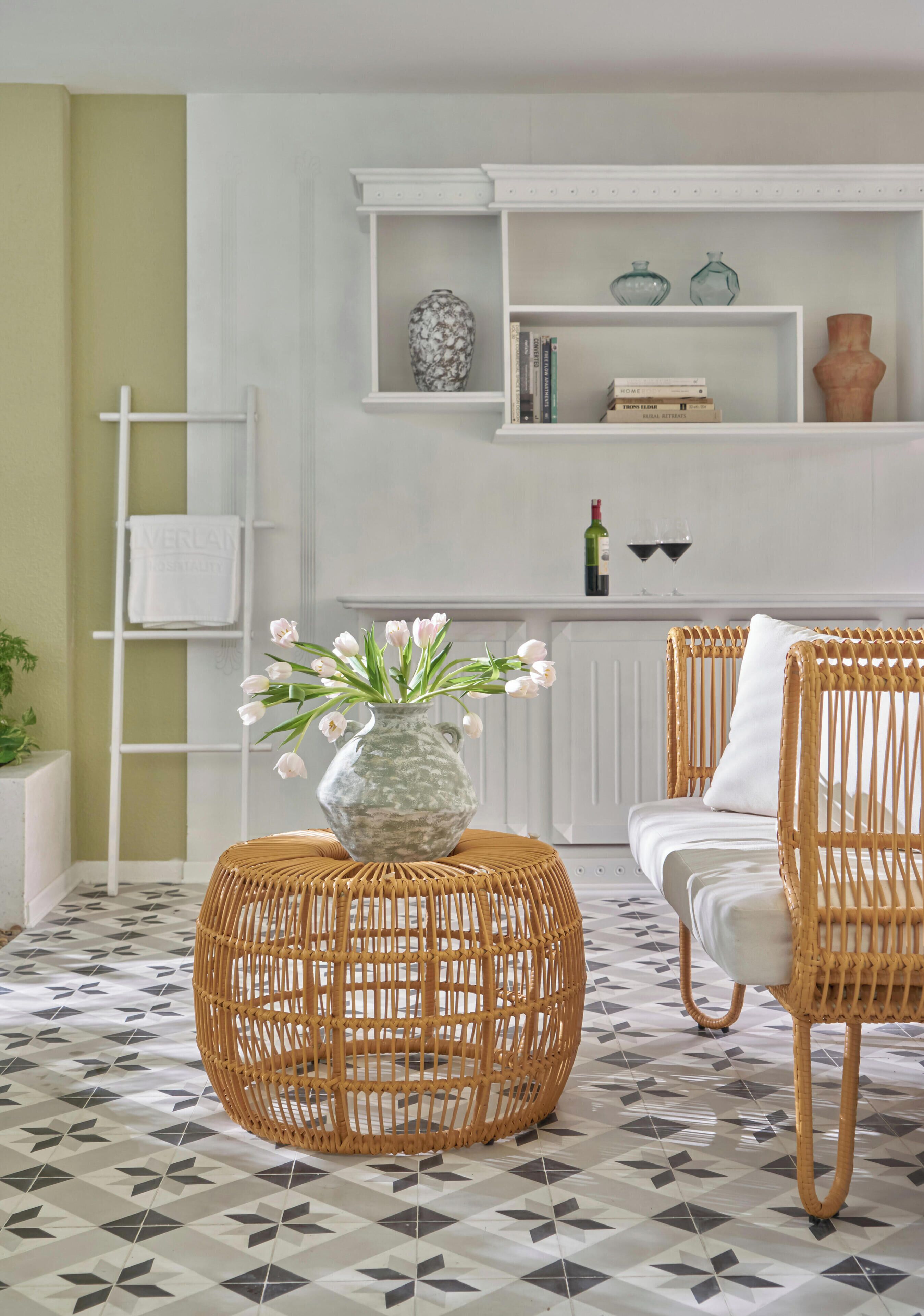 A room with a wicker ottoman, a white sofa, a bookshelf with decorative items, and a patterned floor.