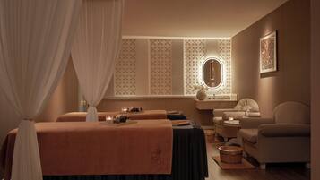 Un cuarto de spa con dos camillas de masaje, un tocador con espejo, una silla y una mesita con velas.