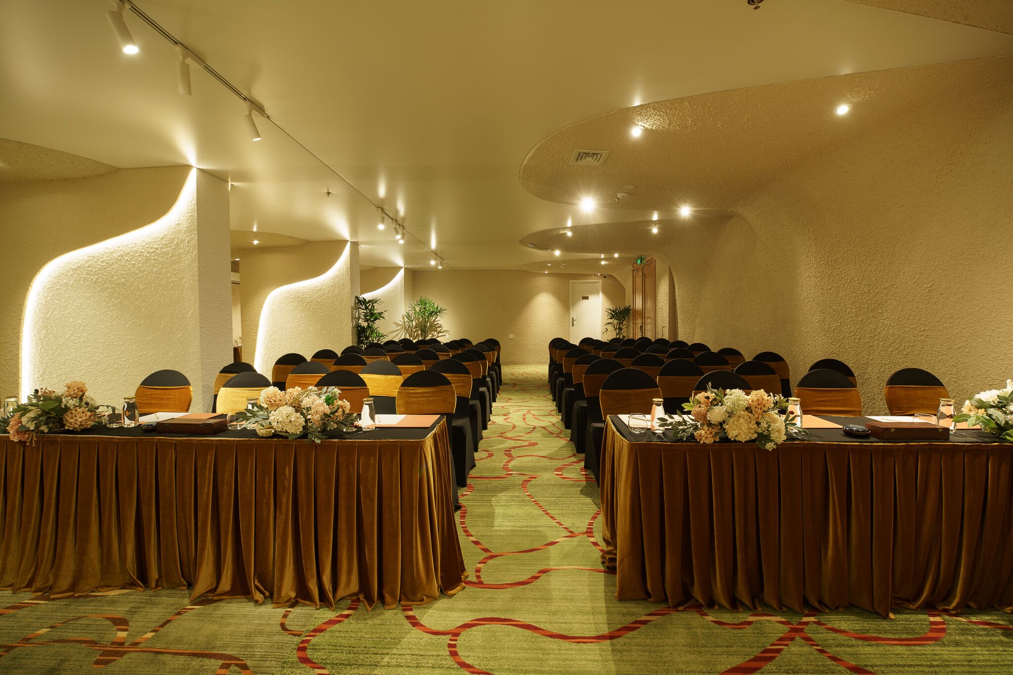 Un salón de conferencias con filas de mesas y sillas, suelo alfombrado e iluminación empotrada en el techo.