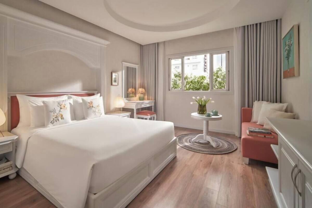 Quarto de hotel com uma cama grande, mesa de cabeceira, abajur, escrivaninha, cadeira e vista para o verde lá fora.