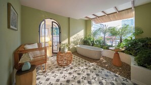 A room with a wicker chair, a small table with a vase, a bathtub, and a view of buildings through a window. - Silverland Mây Hotel (Ho Chi Minh City)