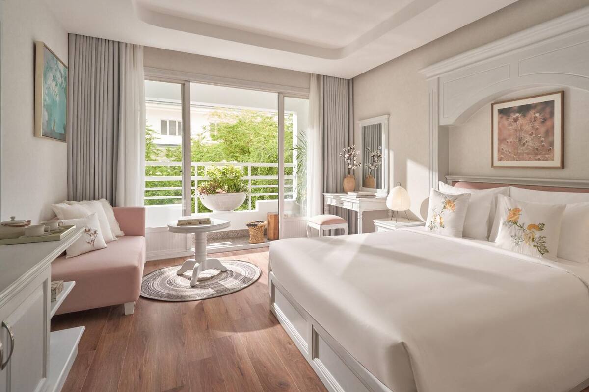 Quarto de hotel com uma cama grande, área de estar com sofá e uma mesinha, escrivaninha com cadeira e vista para o verde lá fora.