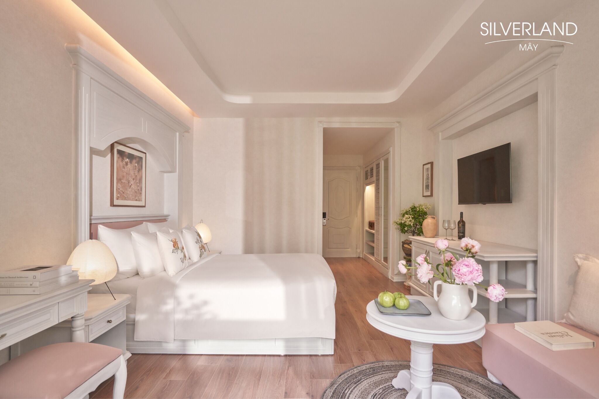 Une chambre d’hôtel avec un grand lit, des tables de chevet, une télévision, une petite table avec des fleurs et une bibliothèque.