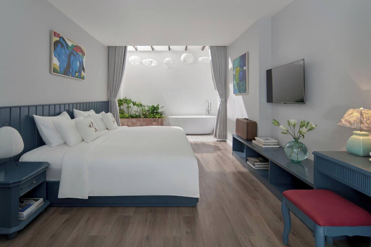 Quarto de hotel moderno com uma cama grande, uma escrivaninha com abajur, televisão e vista para o jardim.