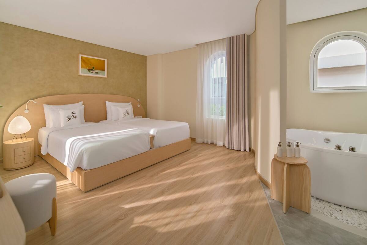 Quarto de hotel com cama de casal, piso de madeira, banheiro com banheira e janela com cortinas.