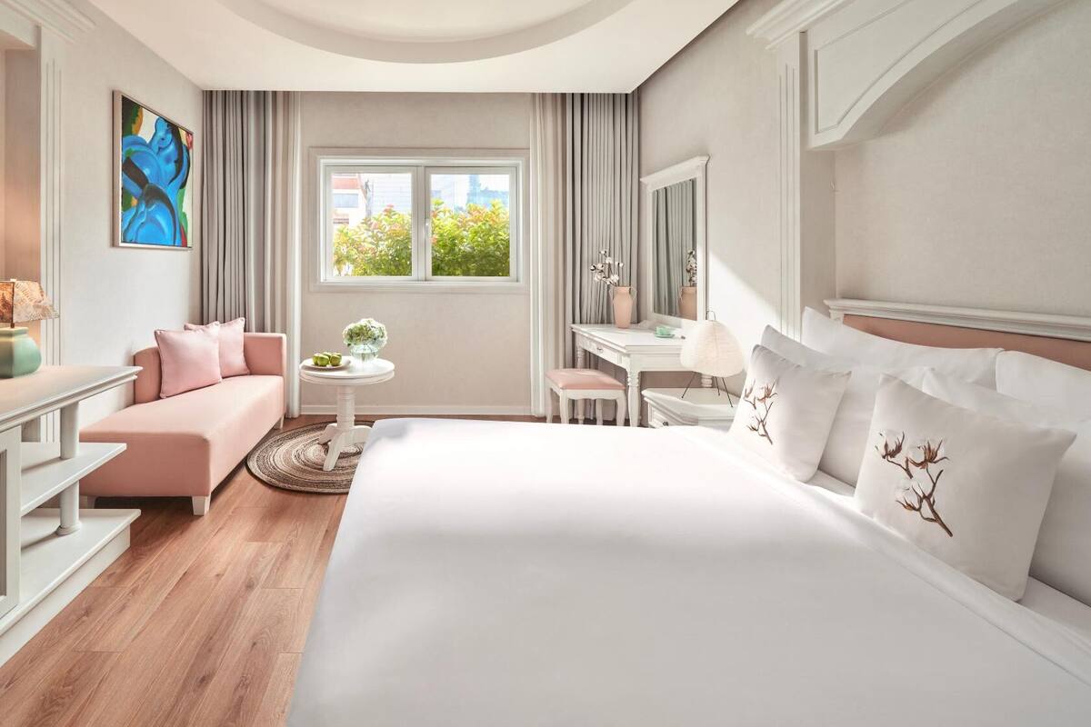 Quarto de hotel com cama, escrivaninha, sofá, televisão e janela com vista para árvores.