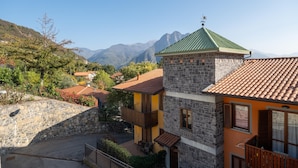Ein Steinhaus mit einem grünen Dach und einem Balkon, umgeben von anderen Gebäuden und Bergen im Hintergrund.