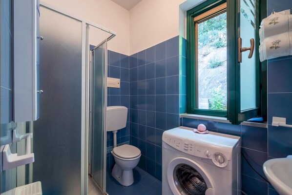 A bathroom with a toilet, a washing machine, and a window with a view of greenery.