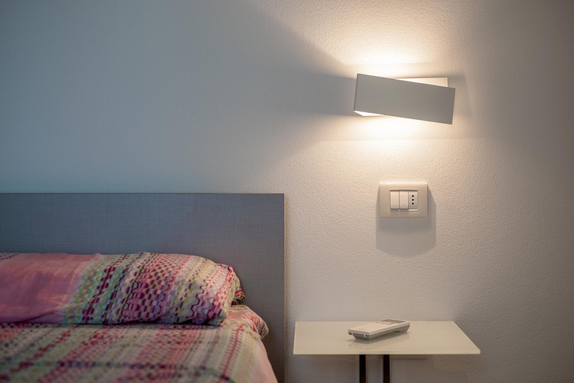 Una cama bien hecha con un edredón estampado, una mesita de noche con un control remoto y una lámpara fijada a la pared.
