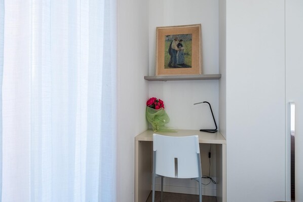 Una habitación pequeña y bien iluminada con un escritorio, una silla, un cuadro enmarcado y un ramo de flores.