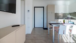 A modern kitchen with white cabinets, a dining table with chairs, a TV mounted on the wall, and a door leading to another room. - Dali in Spiglia With 2 Bedrooms and 1 Bathrooms (Parzanica)