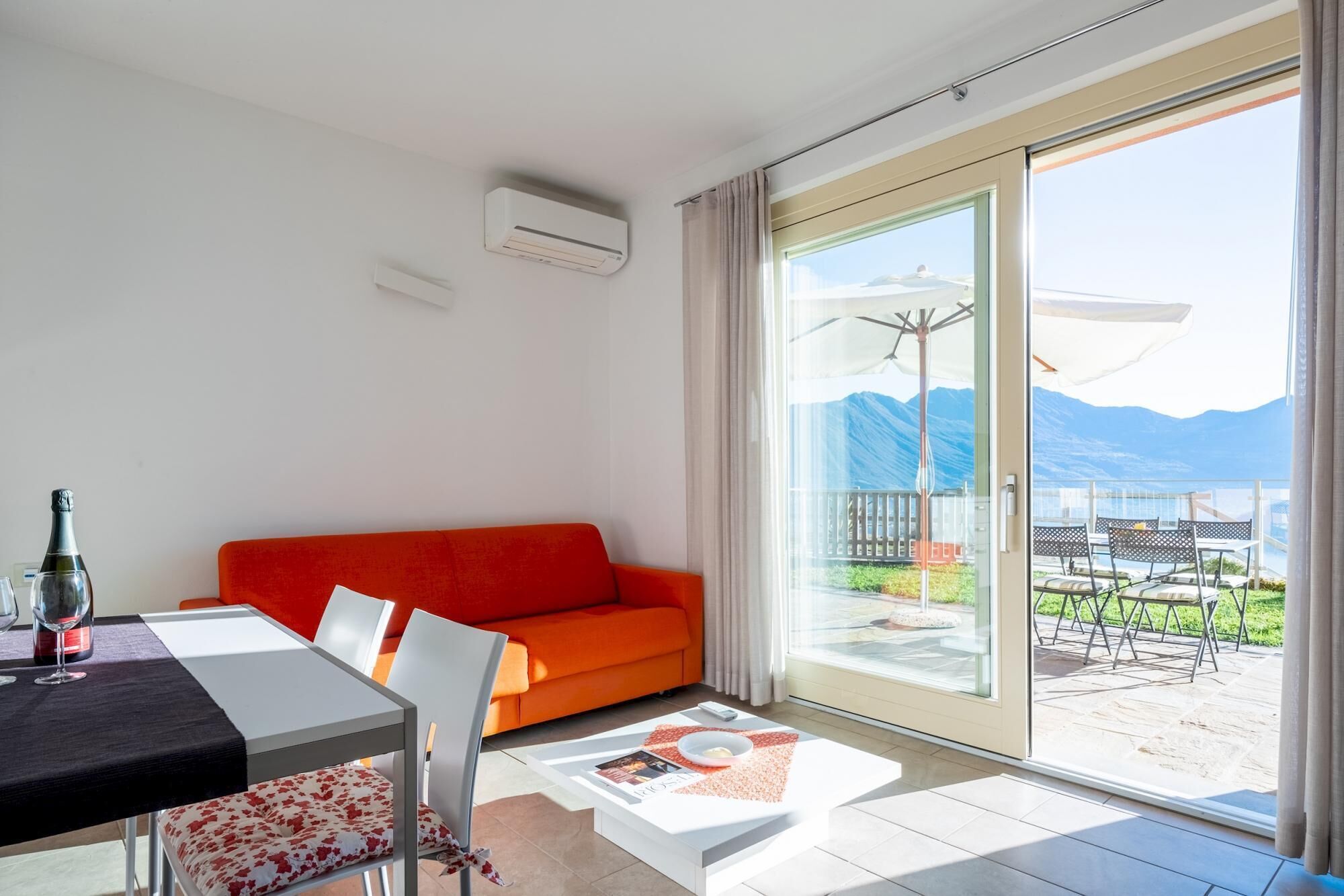 A modern living room with a red sofa, a dining table with chairs, and a large sliding glass door leading to an outdoor patio with a view of mountains.