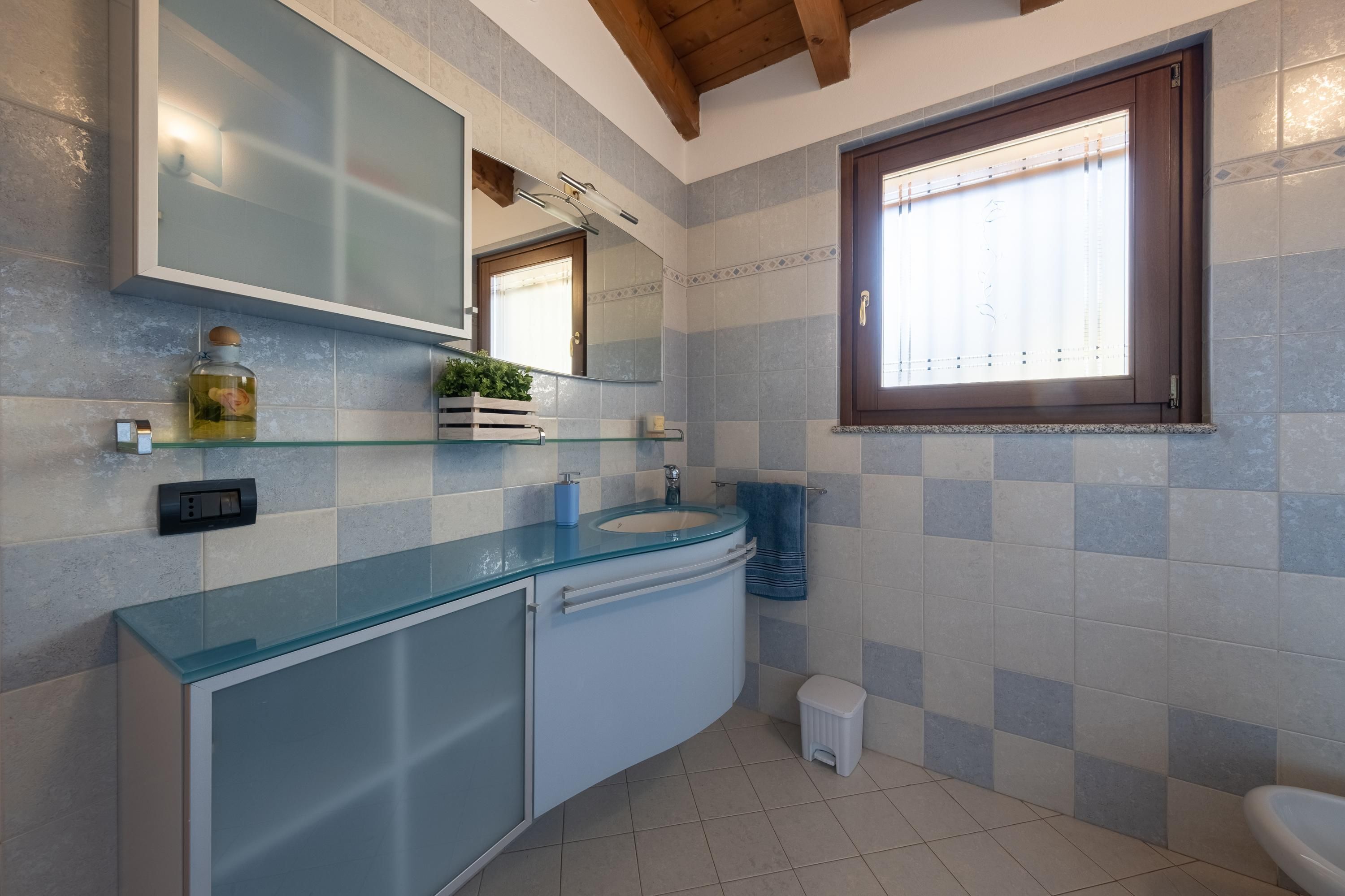 A bathroom with a glass countertop, a sink, a mirror, and a window.