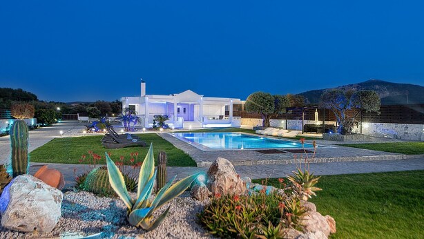 Una casa moderna con piscina, rodeada de un jardín cuidado con diversas plantas y una vista despejada de las montañas al fondo.