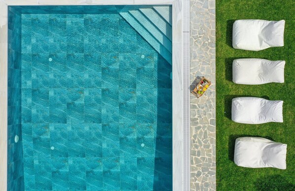 A rectangular pool with clear blue water, surrounded by a stone border and a grassy area with white cushions.