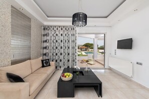 Un salon moderne avec un canapé, une table basse sur laquelle on a disposé des fruits, un lustre et une vue sur une piscine.