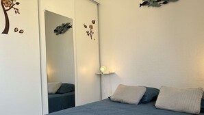 A bedroom with a bed, two pillows, a nightstand with a lamp, and wall decorations including a tree and flowers. - Cozy 1BR Apt with Balcony, A/C, Near Beach and Port - Sleeps 4 (Fleury-Saint-Pierre-la-Mer)