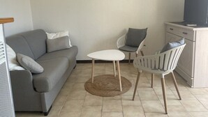 A living room with a grey sofa, a small round table, two chairs, a TV on a cabinet, and decorative wall mirrors. - Cozy 1BR Apt with Balcony, A/C, Near Beach and Port - Sleeps 4 (Fleury-Saint-Pierre-la-Mer)