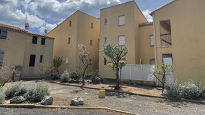 A courtyard with buildings, a small tree, and a hose. - Cozy 1BR Apt with Balcony, A/C, Near Beach and Port - Sleeps 4 (Fleury-Saint-Pierre-la-Mer)