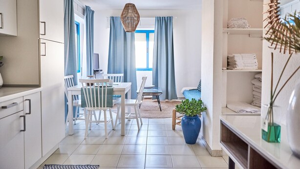 A modern kitchen with white cabinets, a dining area with a table and chairs, a blue rug, and a potted plant. - Marinella in Golfo Aranci (Golfo Aranci)