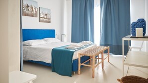 A modern bedroom with a blue headboard, a bed with white linens, a woven bench, a small table with a candle, and two paintings on the wall.