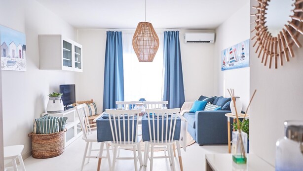 A modern living room with a dining area, featuring a white dining table with chairs, a blue sofa, and a large window with blue curtains. - Cala Coticcio in Golfo Aranci (Golfo Aranci)