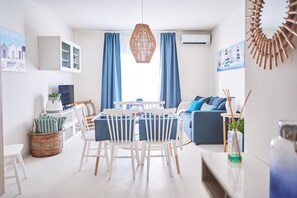A modern living room with a dining area, featuring a white dining table with chairs, a blue sofa, and a large window with blue curtains. - Cala Coticcio in Golfo Aranci (Golfo Aranci)