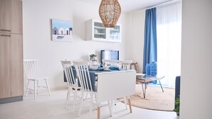 A modern kitchen with white cabinetry, a dining table with chairs, a hanging light fixture, and a window with blue curtains. - Cala Coticcio in Golfo Aranci (Golfo Aranci)