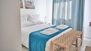 A neatly made bed with white linens and a blue bedspread, a wooden bench, and a wall-mounted painting. - Liscia Ruja in Golfo Aranci (Golfo Aranci)