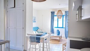 A bright kitchen with white cabinets, a dining area with a blue table and white chairs, and a view of a blue window with curtains. - Liscia Ruja in Golfo Aranci (Golfo Aranci)