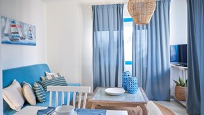 A modern living room with a blue sofa, a dining table with a plate and cups, a wicker chair, and a TV on a shelf. - Liscia Ruja in Golfo Aranci (Golfo Aranci)