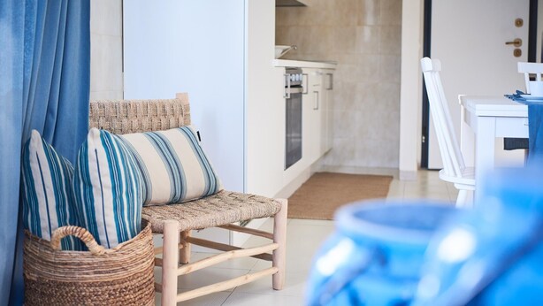 A room with a wicker chair, striped pillows, a woven basket, and a kitchen area in the background. - Liscia Ruja in Golfo Aranci (Golfo Aranci)