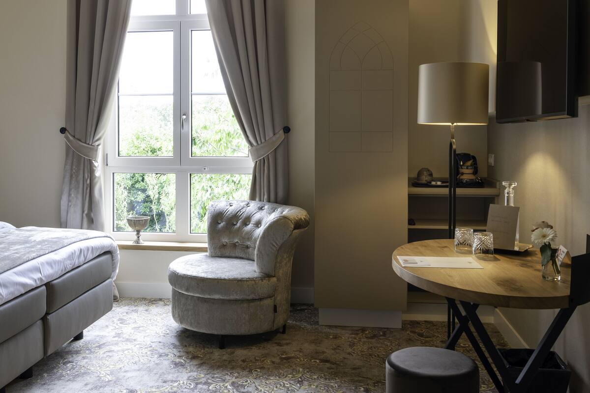 A bedroom with a bed, a tufted armchair, a round wooden table with a lamp, and a window with curtains.
