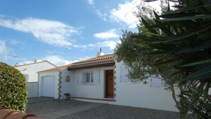 Una casita con techo de tejas, una puerta principal y un ventanal pequeño. Se aprecia un camino de grava y un árbol en primer plano.