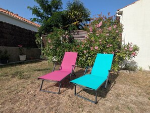 Two outdoor lounge chairs in a garden setting with a white building and a stone wall in the background.