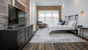 A modern bedroom with a brick wall, a large TV, a bed with white bedding, and a window with a view.