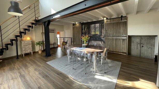 A spacious loft with a high ceiling, exposed beams, a wooden staircase, a dining area with a round table and chairs, and a kitchen with wooden cabinets.