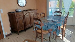 A dining area with a table covered in a blue tablecloth, wooden chairs, a wooden cabinet with a radio, and a tiled floor.