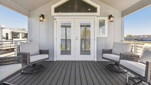 Een overdekt terras met twee banken en een tafel, een witte deur met glaspanelen en een uitzicht op campers op de achtergrond.