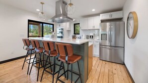 A modern kitchen with a central island, bar stools, stainless steel appliances, and a large window providing a view of the outdoors.