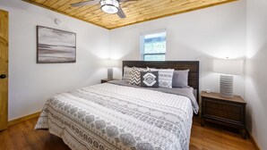 A bedroom with a wooden ceiling, a bed with patterned bedding, two bedside lamps, a window with blinds, and a wall-mounted light fixture. - Welcome Bow- 6 guest/Hot Tub/Location (Broken Bow)