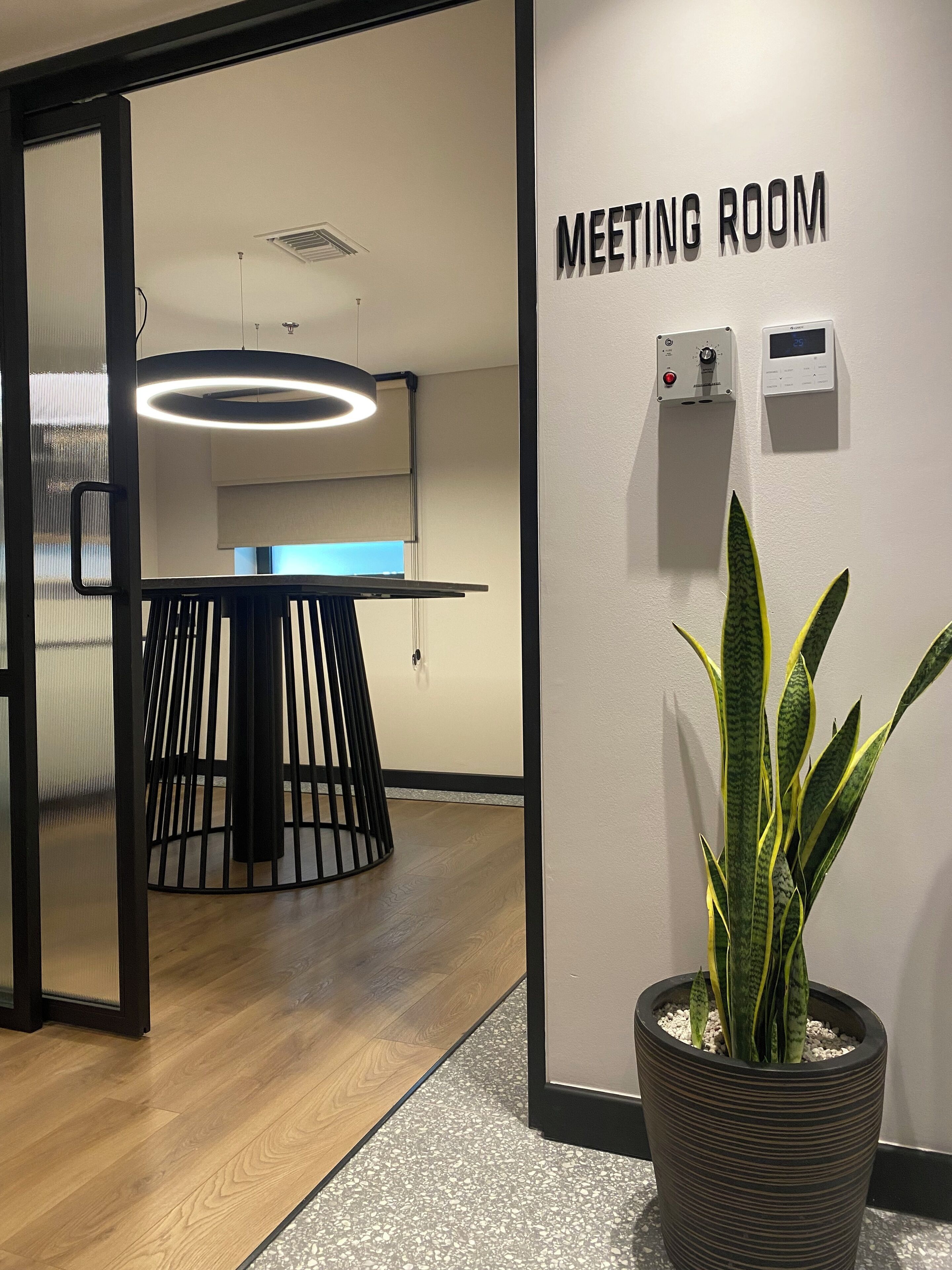A modern meeting room with a wooden floor, a large potted plant, and a sign indicating the room's purpose.