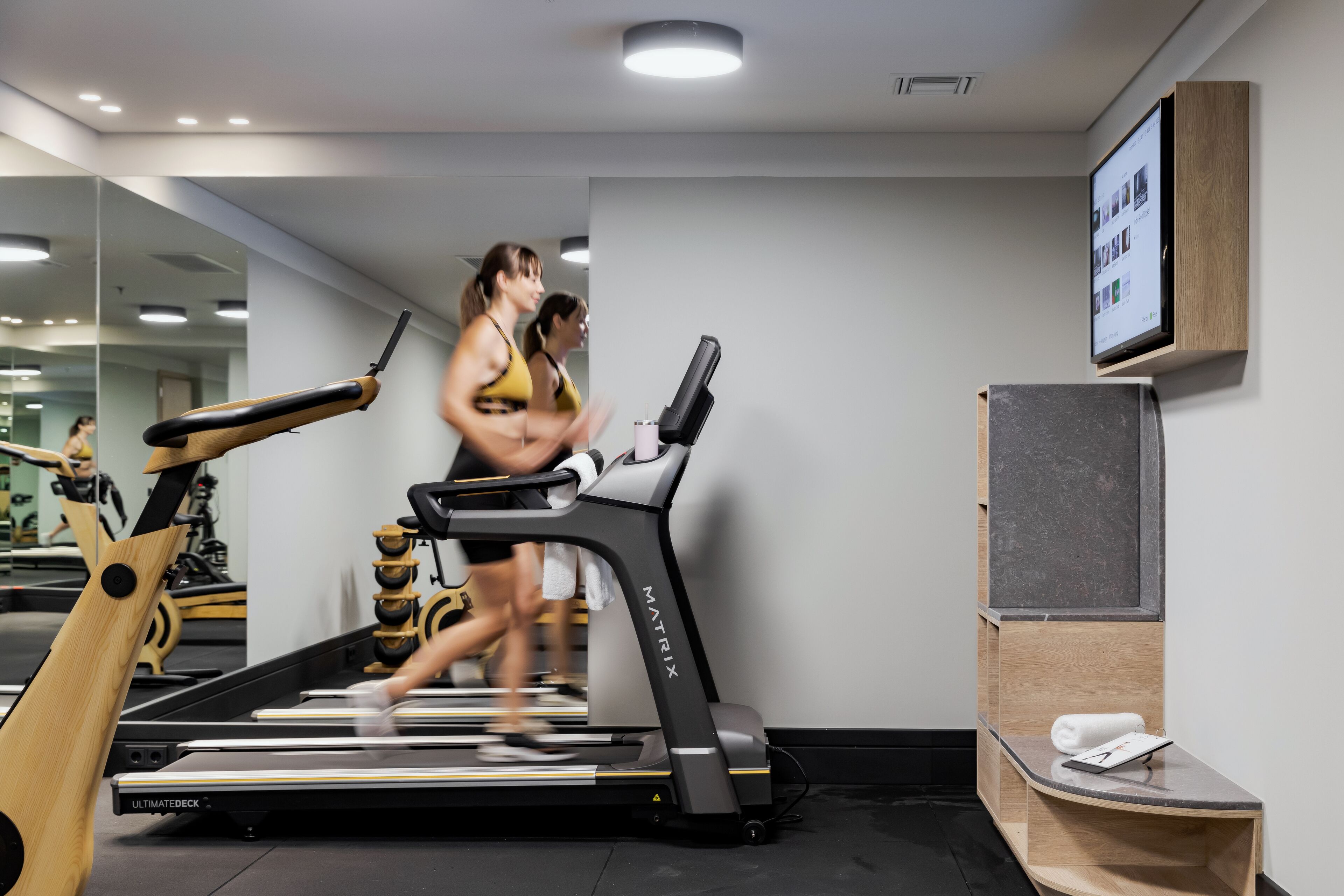 Une salle de sport équipée de tapis de course, d’un appareil d’exercice en bois et d’un écran fixé au mur.