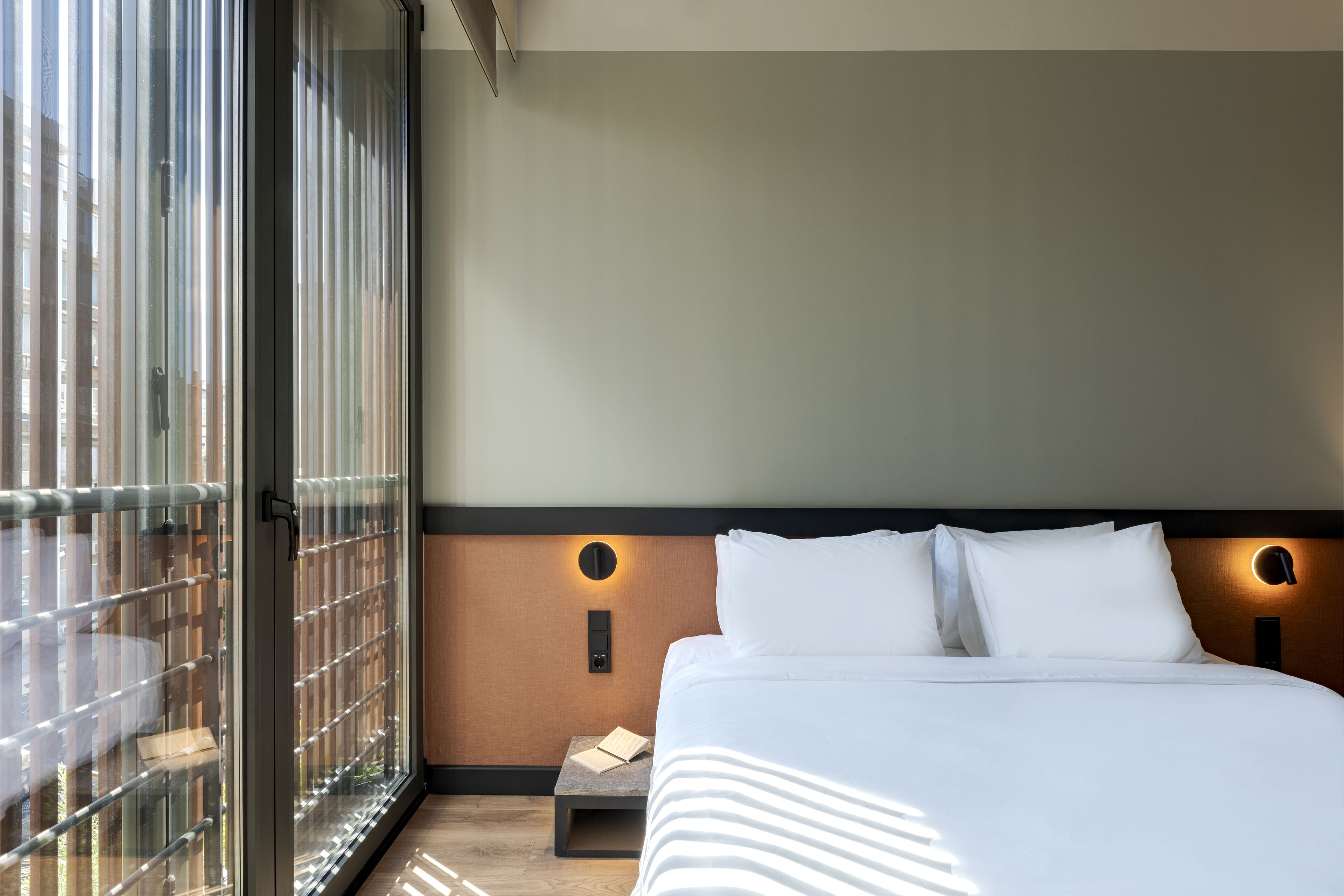 Een hotelkamer met een groot raam, een bed met witte lakens, een nachtkastje en een wandlamp.