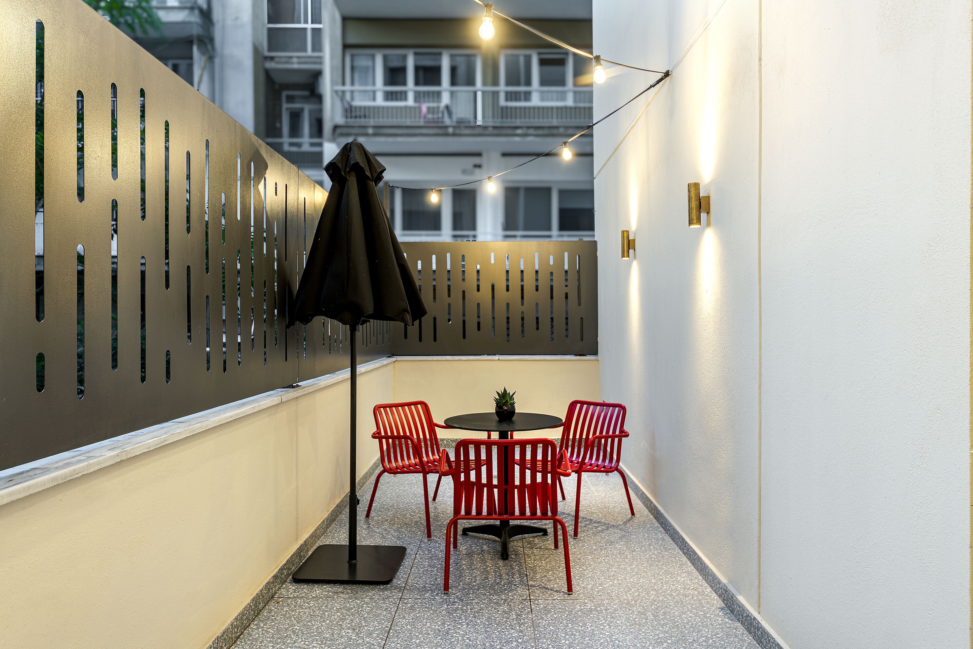 A narrow outdoor corridor with a metal wall featuring vertical slits, a small round table with red chairs, and a black umbrella.